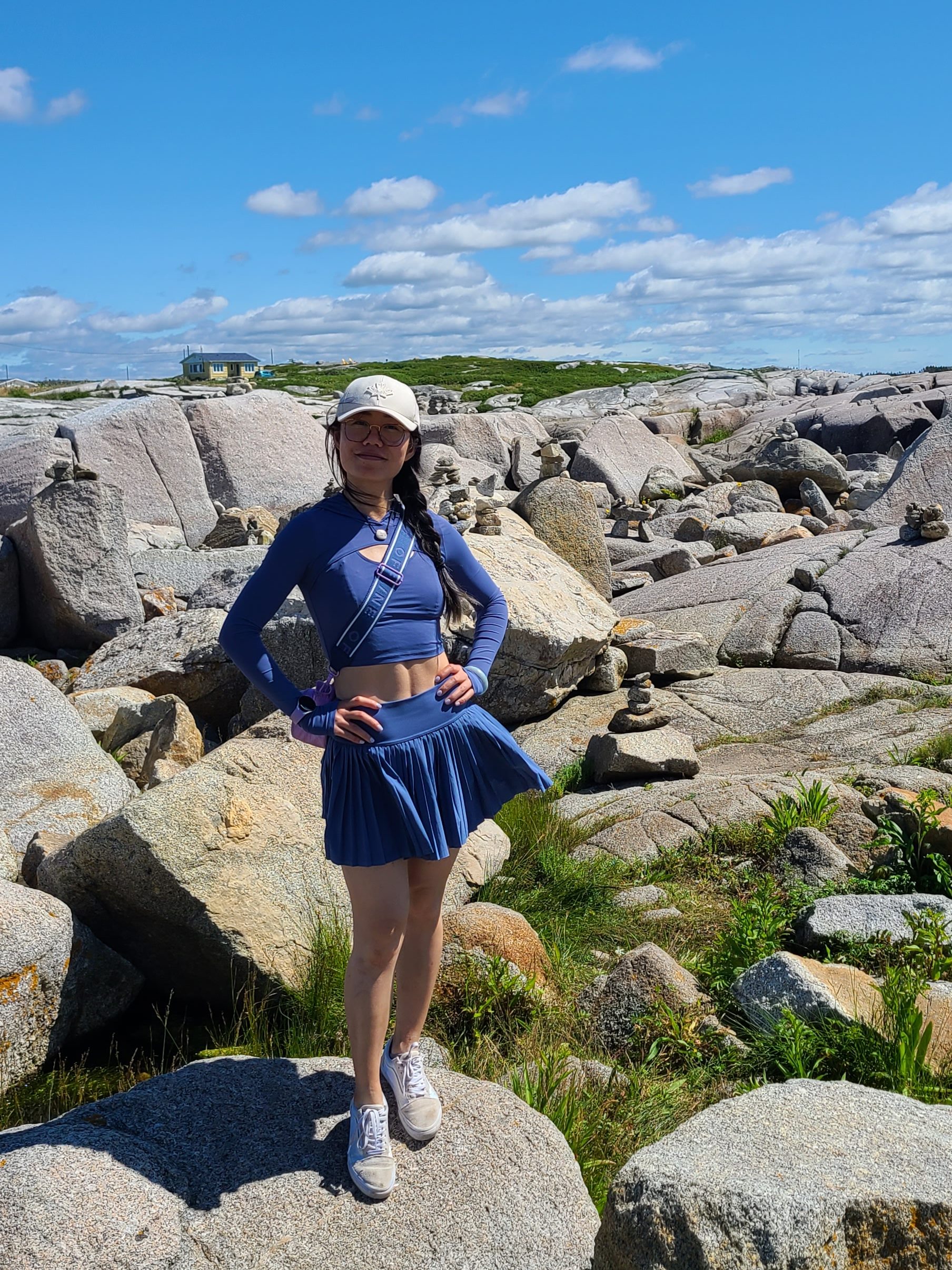 Me standing on the boulders in Peggy's Cove in a matching blue athletic set. The set consist of a form fitting long sleeve crop top and a pleated tennis skirt. My arms are on my hips as I pose for the camera with a clear ab line in my exposed midriff. (lol)